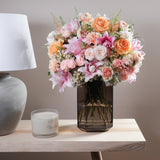 Bouquet of flowers in a vase on a table with a candle and lamp in the background.